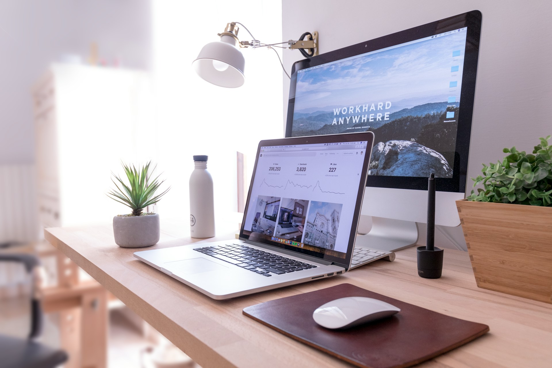 Modern home office setup with a laptop showing analytics and an iMac with 'WORK HARD ANYWHERE' wallpaper, complemented by indoor plants and a stylish desk lamp.